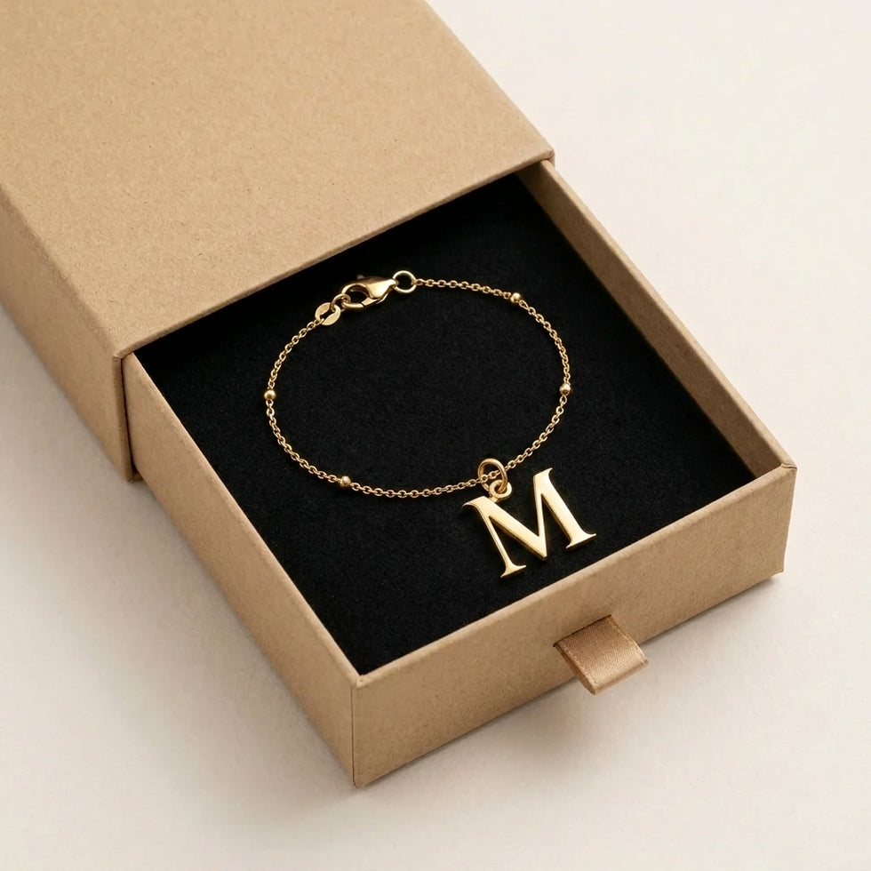 Personalised gold initial bracelet with the letter M in the Mia style, presented in the kraft gift box it is sent in.
