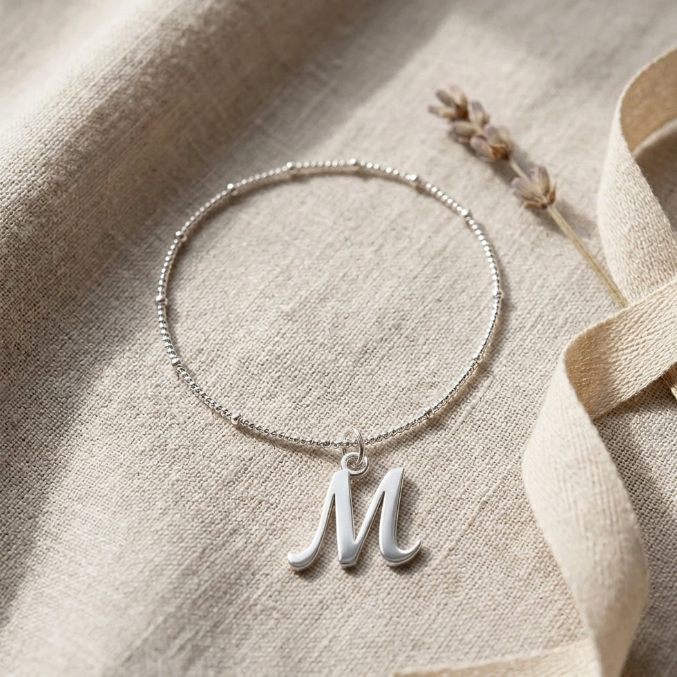 Personalised silver initial bracelet with the letter M in the Bella style, styled on linen with dried flowers.